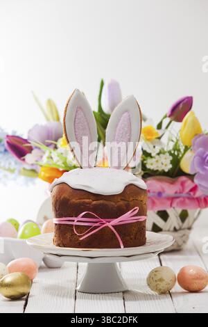 Herrliche Osterkuchen, bedeckt mit weißem Zuckerzusatz und dekoriert mit Hasenohren, auf einem weißen Kuchenständer platziert. Korb mit Blumenstrauß auf Backgro Stockfoto