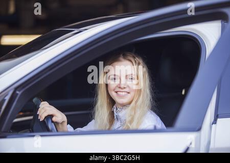 Attraktive junge blonde Frau, die im weißen Auto sitzt, lächelt und in die Kamera schaut Stockfoto