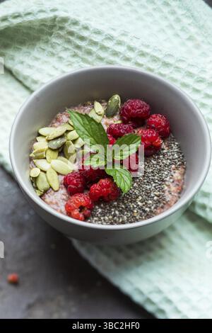 Schüssel Haferbrei mit frischen Himbeeren, Kürbiskernen und Chiasamen, serviert auf Leinen Küchentuch über graue Oberfläche. Rustikales Frühstücksmotiv. Oben vi Stockfoto