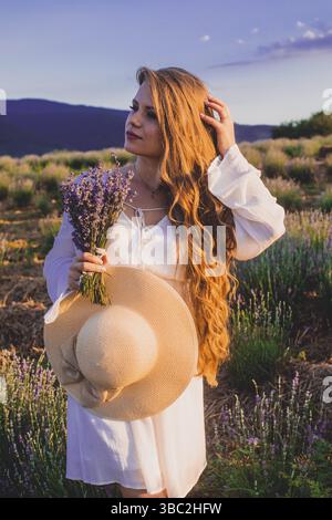 Porträt einer schönen blonden Frau, die zwischen den Plantagen von Lavendel steht. Mädchen in einem weißen Sundress gekleidet und hält sbunch Lavendel Stockfoto