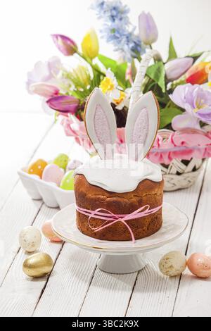 Herrliche Osterkuchen, bedeckt mit weißem Zuckerzusatz und dekoriert mit Hasenohren, auf einem weißen Kuchenständer platziert. Korb mit Blumenstrauß auf Backgro Stockfoto