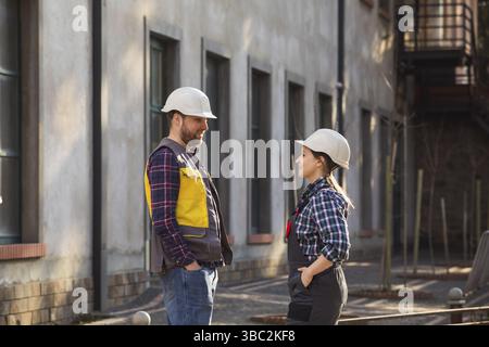 Zwei junge Fabrikangestellte, die sich während der Mittagspause im Freien unterhalten. Frau und Mann in Schutzhelmen, die sich auf dem Werksgelände anschauen. Romantisch Stockfoto