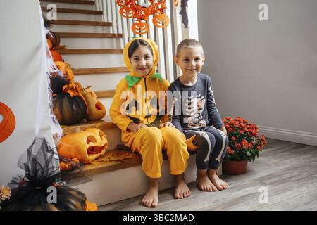 Porträt von fröhlichen Brüdern und Schwestern in halloween-Kostümen, die zu Hause auf verzierten Holztreppen sitzen. Die Kinder sind auf die halloween-Party vorbereitet Stockfoto