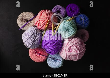 Von oben sehen Sie einen Haufen Plüsch- und Textilgarnbälle mit Stricknadeln und Haken auf dem schwarzen. Hobby-Still-Leben Stockfoto