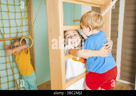 In der Nähe der Brüder und Schwester spielen in einem Holz- Startseite Sport Komplex. Mädchen weit lächelnd, ihr Bruder, ihm helfen, Holz- Bars zu klettern Stockfoto