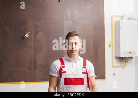 Der hübsche junge Klempner im Overall steht an der Wand. Der Mann ist im Heizungsraum, der gerade renoviert wird Stockfoto