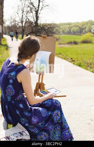 Junge Künstlerin, die im Frühjahr an der Malerei im Freien arbeitet. Der Künstler malt mit Aquarell auf dem Pult Stockfoto