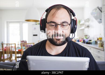 Erwachsene junge Mann im Headset sprechen von Webcam Blick auf Tabelt Computer zu Hause. Gespräche per Videoanruf mit Küche Zuhause Stockfoto