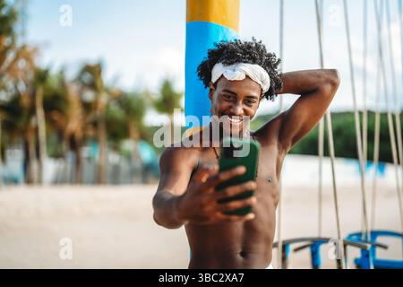 Stilvoller junger schwarzer Mann, der einen sonnigen Strandtag genießt, während er ein Selfie mit seinem Smartphone macht Stockfoto