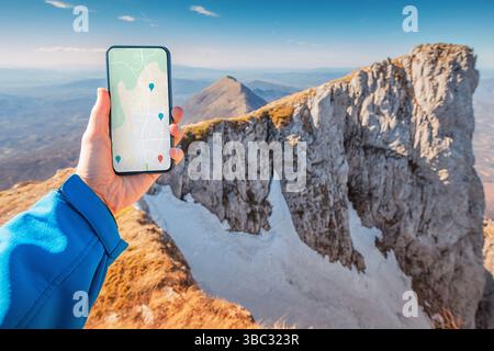 Handheld-Smartphone, das Navigations-App mit Karten- und Positionsnadeln in Berglandschaft anzeigt Stockfoto