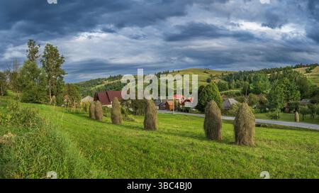 Malerische ländliche Landschaft in den Karpaten mit Heuhaufen auf grünen Hügeln und Bäumen unter einem dramatischen Himmel. Friedliche Aussicht auf die Landschaft Stockfoto