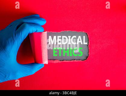 Medizinethik-Symbol. Konzeptworte medizinische Ethik auf wunderschönem grauem Papier. Arzthand in blauem Medizinhandschuh. Schöner roter Hintergrund. Gesundes Leben Stockfoto