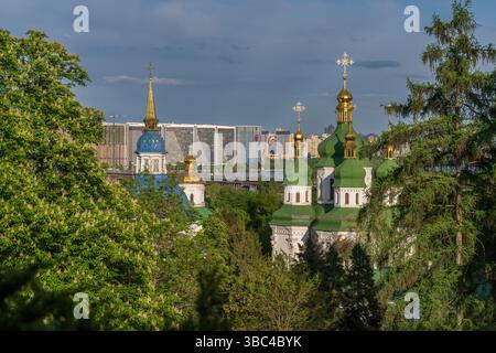 Malerischer Blick auf das Vydubychi-Kloster in Kiew mit goldenen Kuppeln und grünen Dächern vor der modernen Skyline des linken Dnipro-Ufers Stockfoto