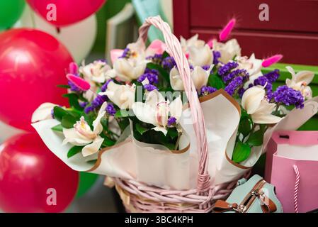 Wunderschönes Arrangement aus weißen und lila Blumen in einem rosafarbenen Korb, umgeben von bunten Ballons und Geschenktüten. Stockfoto