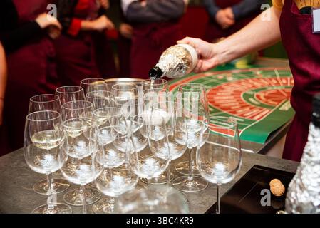 Die Person gießt während einer Party oder Veranstaltung Champagner in mehrere Gläser, mit Personen und einer Matte mit Roulette-Motiv im Hintergrund. Stockfoto
