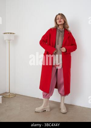 Frau in stilvollem rotem Mantel, beigefarbenen Stiefeln, rosa Hose und braunem Oberteil steht drinnen auf Betonboden, neben einer eleganten Lampe mit einem weißen Farbton, Hände in Stockfoto