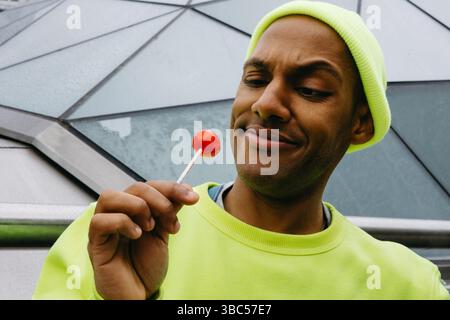Junger Mann, der einen roten Lutscher in urbaner Umgebung amüsiert Stockfoto
