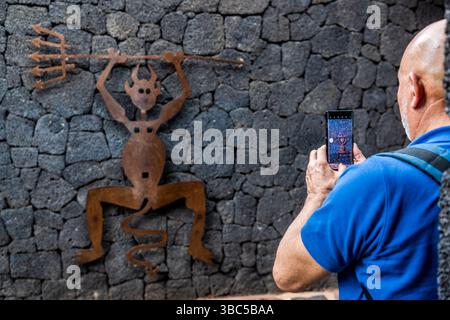 Ein Mann fotografiert die Metallskulptur eines Dämons auf einer Steinmauer im Timanfaya-Nationalpark. Die Figur ist als El Diablo bekannt. Sie wurde vom Künstler César Manrique entworfen und dient noch heute als Emblem des Parks. Stockfoto