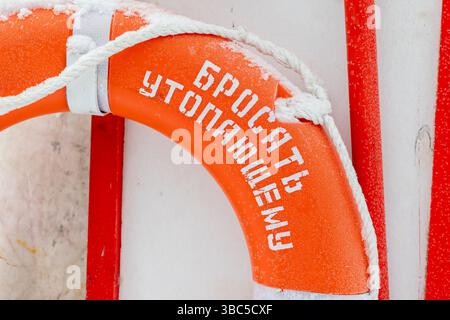 Ein leuchtend orangefarbener, schneebedeckter Rettungsschirm, der die Sicherheitsausrüstung für den Seeverkehr unter winterlichen Bedingungen betont. Der Text auf Russisch ist übersetzt: Werfen zu a d Stockfoto