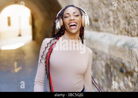 Eine fröhliche junge Frau, die Kopfhörer trägt, während sie fröhlich Musik in einer lebhaften urbanen Umgebung genießt Stockfoto