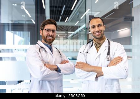 Zwei lächelnde Ärzte in weißen Mänteln und Stethoskopen posieren selbstbewusst mit gekreuzten Armen, bereit für die Konsultation. Stockfoto