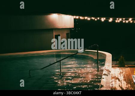 Eine faszinierende Nachtszene zeigt einen Pool mit glitzerndem Wasser unter einer Girlande. Ein Pool, der von sanften Lichtern und einer Reihe dekorativer Glühbirnen beleuchtet wird. Stockfoto
