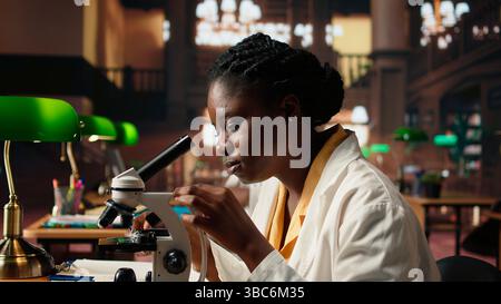 afroamerikaner studiert medizinische Wissenschaft in einer Universitätsbibliothek und erforscht Viren mit einem Mikroskop. Mädchen arbeitet an der Prüfungsvorbereitung für den Erfolg einer wissenschaftlichen Karriere. Kamera A.. Stockfoto
