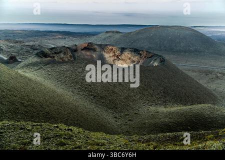 Ein Krater und eine Straße schneiden sich durch die vulkanische Landschaft entlang der Ruta de los Volcanes auf Lanzarote, Yaiza, Kanarische Inseln, Spanien. Die Route verläuft durch den Timanfaya-Nationalpark, Spaniens einzigen rein geologischen Nationalpark. Stockfoto