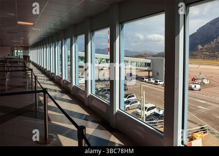 Langer Flughafenkorridor, mit Fenstern gesäumt, von denen aus man auf Asphalt und geparkte Fahrzeuge blicken kann. Blick von innen auf den Flughafen Palermo mit großen Fenstern mit Blick auf den Flughafen Palermo Stockfoto