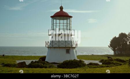 Weißer Leuchtturm mit roter Spitze auf grüner Wiese am Meer unter blauem Himmel Stockfoto