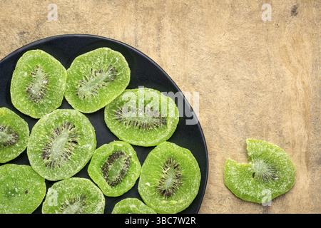 Scheiben getrockneter Kiwis auf einer schwarzen Platte gegen strukturiertes, handgefertigtes Rindenpapier mit Kopierraum Stockfoto