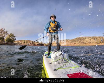 Senior man paddelt ein aufblasbares Stand-Up-Paddleboard gegen Gegenwind auf dem Bergsee - Horsetooth Reservoir, Colorado, in herbstlicher Landschaft Stockfoto