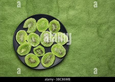 Scheiben getrockneter Kiwis auf einer schwarzen Platte gegen strukturiertes, handgefertigtes Rindenpapier mit Kopierraum Stockfoto