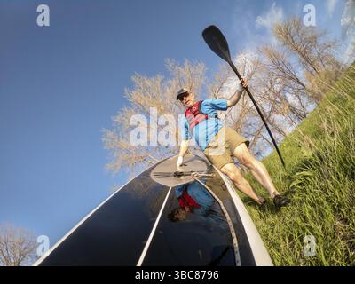 Ein Senior Paddler mit aufblasbarer Schwimmweste und Paddelhandschuhen startet sein Stand Up Paddleboard auf einem See, POV-Bild mit Action-Kamera breit A Stockfoto