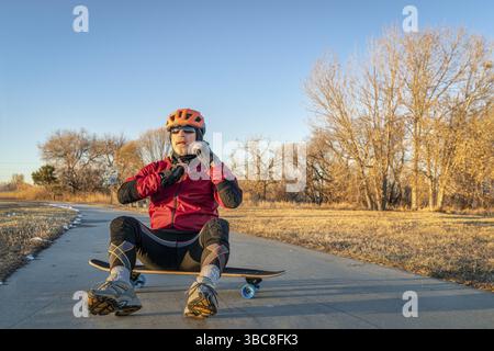 Senior man sitzt auf einem Skateboard und Putting Helm, einem asphaltierten Radweg in der Winterlandschaft im nördlichen Colorado - Boyd Lake State Park Stockfoto