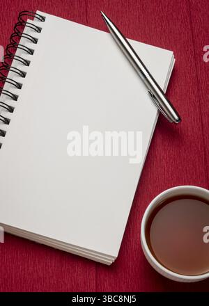 Leeres spiralförmiges Kunstskizzenbuch mit einer Tasse Tee gegen strukturiertes rotes Krepppapier, Arbeitsplatzkonzept Stockfoto