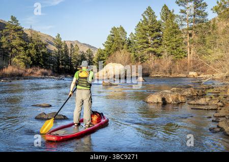 Aufblasbares Stand-Up-Paddleboard für ältere Männer durch den Steingarten auf einem Bergfluss - Poudre River in Colorado in Frühlingslandschaft mit niedrigem Wasser Stockfoto