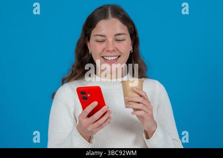 Glückliche kaukasische Frau, die Kaffeetasse und Smartphone hält, lächelt über blauem Hintergrund. Stockfoto