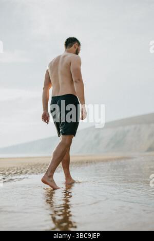 Ein Mann läuft in seinen Shorts am Strand. Das Wasser ist ruhig und der Himmel ist bewölkt Stockfoto