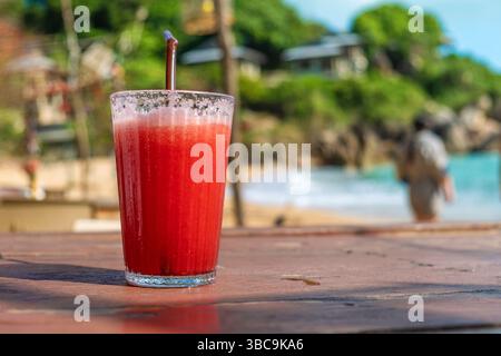 Erfrischender Smoothie mit roter Wassermelone im Glas mit einem Strohhalm auf einem Holztisch im Strandcafe am Meer, Samui Island, Thailand. Kalter tropischer Fruchtsaft Stockfoto