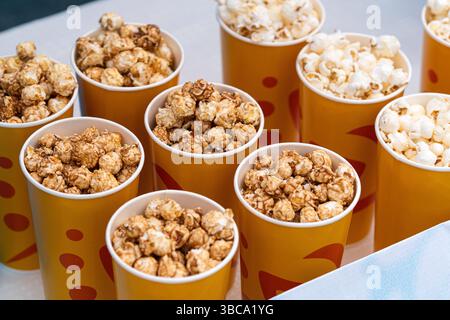 Pappbecher mit Popcorn-Snack auf dem Tisch. Köstliches süßes, karamellbraunes Popcorn. In einer gelb-orangen Glasnahmebildung. Kisten mit Popcorn im Verkauf auf dem C Stockfoto