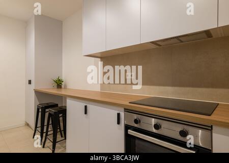 Blick auf eine moderne Küche mit Holztheke, weißen Schränken, Backofen, schwarzen Hockern und kompaktem Layout Stockfoto