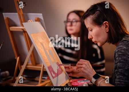 Zwei Frauen konzentrierten sich auf ihre Leinwandkunst während einer entspannten Indoor-Malerei-Sitzung mit Vorräten über den Tisch verteilt. Stockfoto
