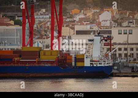 Frachtschiff beladen mit bunten Containern im Hafen von Lissabon, roten Kränen und pulsierenden Gebäuden der Stadt am sonnigen Abend. Dynamische Wohn- und Industrieszene durch Uferpromenade, städtische Skyline, Be- und Entladen, ruhiges Wasser, Handel, Industrie Stockfoto