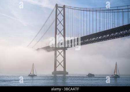 Dramatische Aussicht auf die berühmte Brücke von Lissabon 25 de Abril im Morgennebel an sonnigen Tagen, mit Segelbooten und Motorbooten auf dem ruhigen Fluss Tejo, Portugal. Neblige Atmosphäre, ruhige, geheimnisvolle Szene, perfekt für Reisen, Stadtmotive, Aufhängungsbrücke. Stockfoto