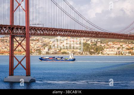 Farbenfrohe Landschaft mit blauem Containerschiff auf dem Tejo unter der berühmten Brücke 25 de Abril oder 25. April in Lissabon, Portugal. Berühmtes Eisenwerk der roten Hängebrücke, pulsierende Stadtlandschaft, städtische Architektur, Seeverkehr, sonniger Tag, bewölkter Himmel Stockfoto