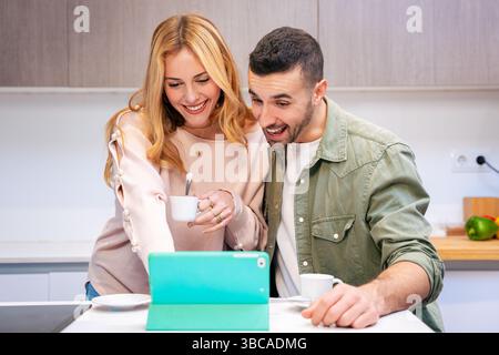 Glückliches Paar, das ein Tablet in der Küche zusammen verwendet, während es Kaffee genießt und lächelt Stockfoto