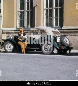 Eine Frau bei einem Sportwagen. Der Wagen ist ein englisches MG TD-Modell, das zwischen 1950 und 1953 hergestellt wurde. Dieses Modell war besonders für schwedische Kunden geeignet, da es mit dem Lenkrad auf der linken Seite gebaut werden konnte, was den damaligen schwedischen Fahrkonventionen entsprach. Insgesamt wurden 29.664 Einheiten des MG TD hergestellt. Sie sitzt auf dem Auto und liest die Frauenzeitschrift Femina. Sie trägt einen Blazer und eine braune Hose. Schweden der 1960er Jahre. Weitere Fakten zum MG TD der MG TD, Teil der MG T-Type-Serie, war ein zweisitziger Roadster, der für sein klassisches britisches Design bekannt ist Stockfoto