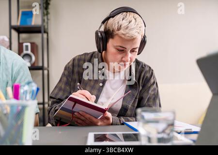 Lässiger Millennials-Profi, der Kopfhörer trägt und Ideen in einem gestreiften Notebook an einem modernen Coworking-Schreibtisch schreibt, Konzept von Fokus, Studium und Crea Stockfoto