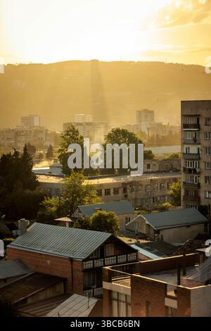 Goldenes Sonnenlicht strömt durch Wolken über die Stadtlandschaft von Tiflis und wirft bei Sonnenuntergang dramatische Lichtstrahlen über Wohngebäude und Bäume. Warm, bei Stockfoto